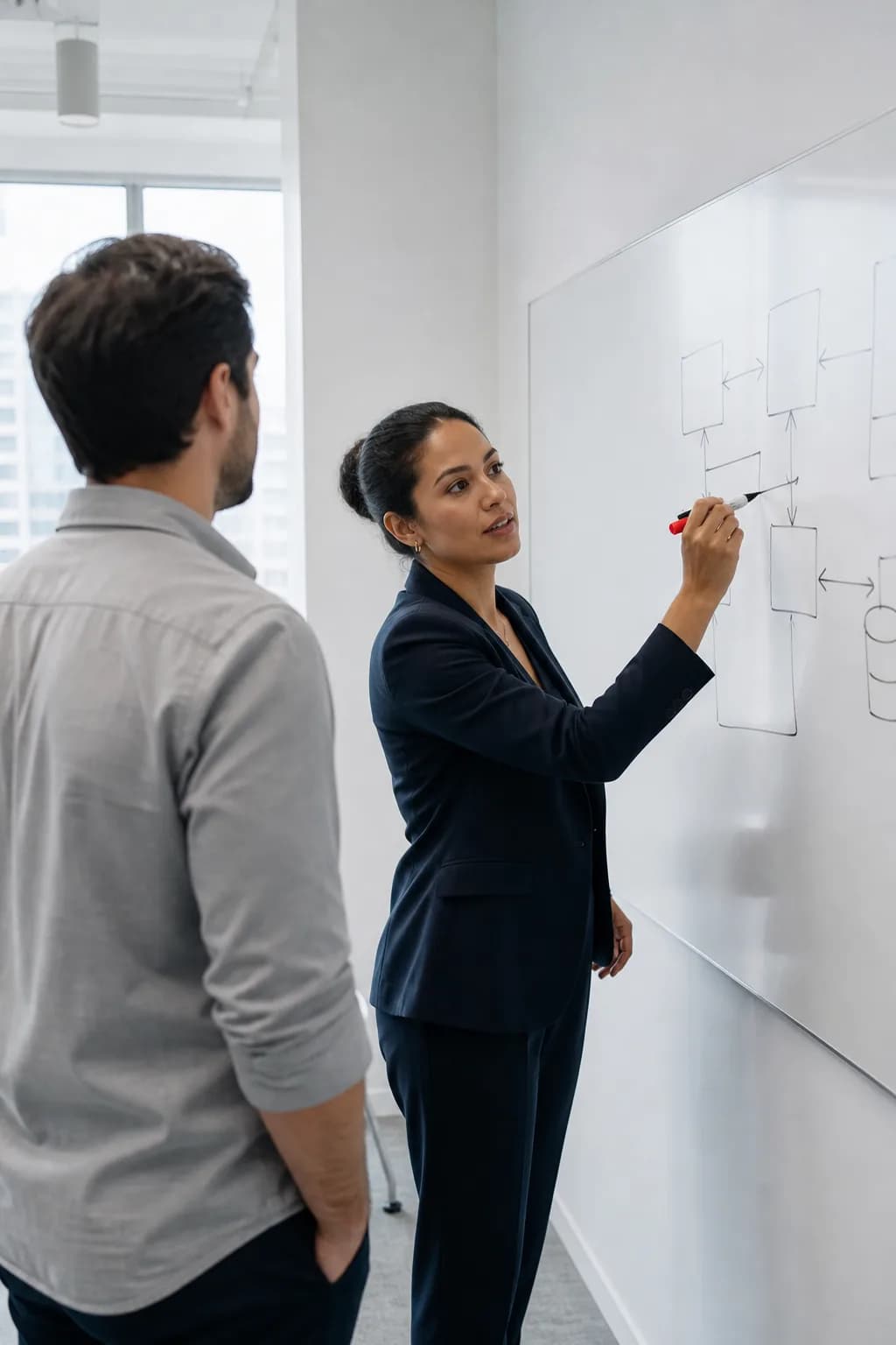 A consultant sketches a system diagram on a whiteboard while a colleague watches, in a bright office